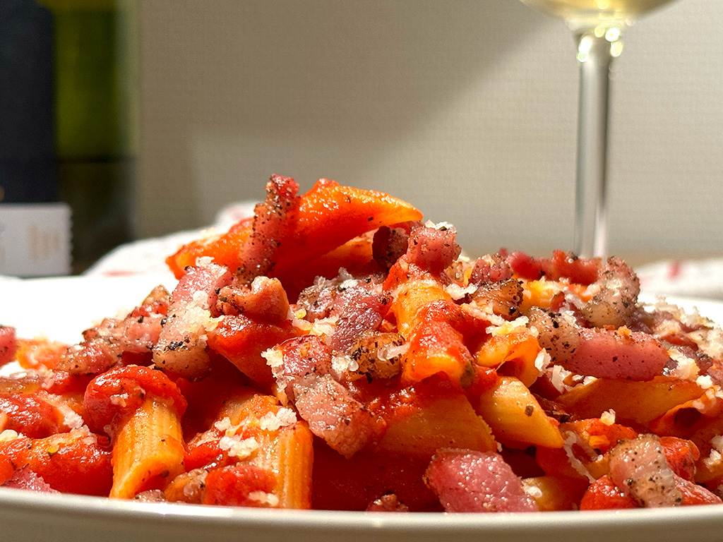 A plate of penne with bacon with wine in the background